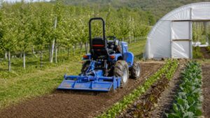 Illustration of a mini tractor working with a rotary tiller near orchard rows and a greenhouse